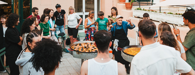 Viele Erwachsene stehen um einen Koch mit zwei Paellapfannen im Innenhof der Sprachschule Barcelona