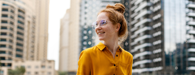 Bildungsurlaub Englisch in Dubai Eine junge Frau mit Brille und orangefarbener Bluse sitzt in Dubai vor Hochhäusern