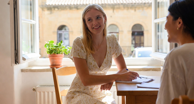 Lernen in der Sonne bei LISA! Sprachreisen Eine junge Frau mit blonden Haaren sitzt in einem Klassenzimmer bei offenem Fenster in mediterraner Umgebung