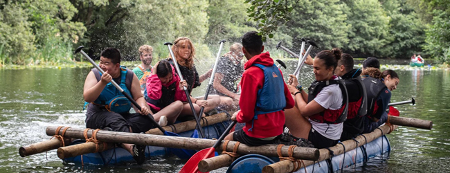 Boostfahrt auf einem See als Aktivität bei Adventure England Schüler sitzen in einer Art Boot aus Fässern und paddeln über einen See bei Adventure England